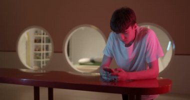 Millenial Guy Smartphone 'unu kullanarak, Neon Lighting ve Futuristik Oda Arkaplanı' nda Rack Indoor 'a yaslanırken ekranı Scrooling the Screen' i kullanarak. Geniş Açı Video.