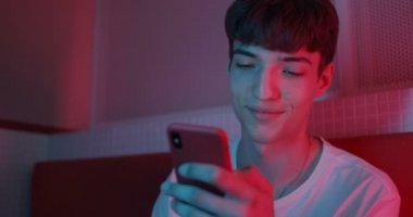 Beyaz tişörtlü Millenial Boy 'un Smartphone' unu kullanırken yakın plan çekimi. Neon Club Işıklandırma Arkaplanındaki koltukta otururken Ekranda Sürünen ve Gülümseyen Adam.