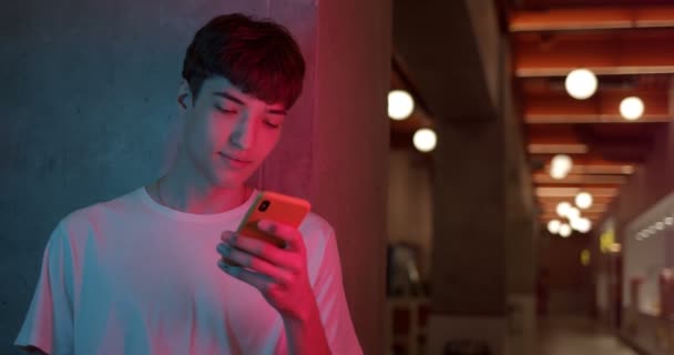 Young Stylish Millenial Boy in White T-Shirt Usando il suo smartphone per chattare, digitando messaggi mentre si trova in piedi sullo sfondo futuristico delle luci del Neon Club. Chiudere colpo . — Video Stock