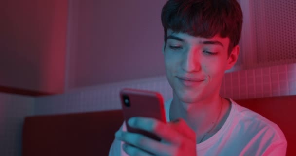 Close Up Shot of Young Stylish Millenial Boy in White T-Shirt Usando il suo smartphone. Uomo Scrooling lo schermo e sorridente mentre seduto sul divano a Neon Club Illuminazione sfondo . — Video Stock