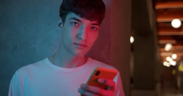 Portrait of Young Stylish Millenial Boy in White T-Shirt Holding his Smartphone and Looking straight to Camera. Man Standing at Futuristic Neon Club Lights Background. Close Up Shot. — Stock Video