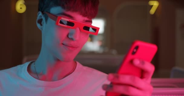 Close Up Shot of Stylish Cool Millenial Guy w modnych okularach Korzystanie z jego nowoczesny smartfon i uśmiechnięty, Młody człowiek siedzi w domu z Neon Lighting w tle. — Wideo stockowe