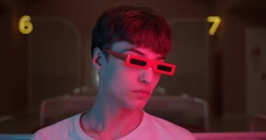 Havalı Millenial Guy 'ın Portresi Moda Partisinin Gözlükleri Dönüyor ve Doğrudan Kameraya Bakıyor Fütürist Neon Işıklandırma Numaralarıyla Arka planda. Yakın çekim.