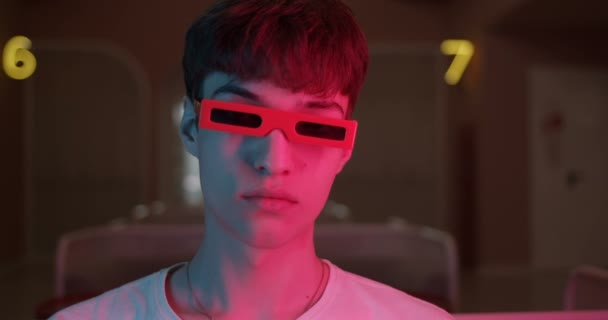 Портрет молодого парня в белой футболке в смешных 3D-очках, смотрящего прямо в камеру с футуристическими неоновыми номерами освещения на заднем плане. Закрыть выстрел . — стоковое видео