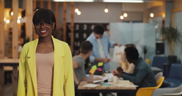 Femme d'affaires afro-américaine debout dans le bureau moderne, souriant et regardant dans la caméra. Les travailleurs sur le fond flou . — Video