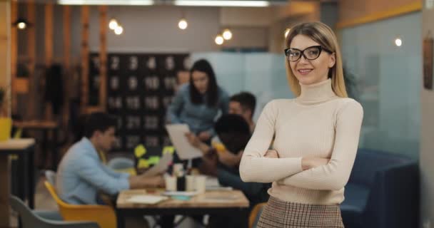 Ritratto di giovane donna d'affari di successo con occhiali alla moda guardando la fotocamera in piedi nello spazio di lavoro dell'ufficio. Persone che lavorano sullo sfondo sfocato . — Video Stock