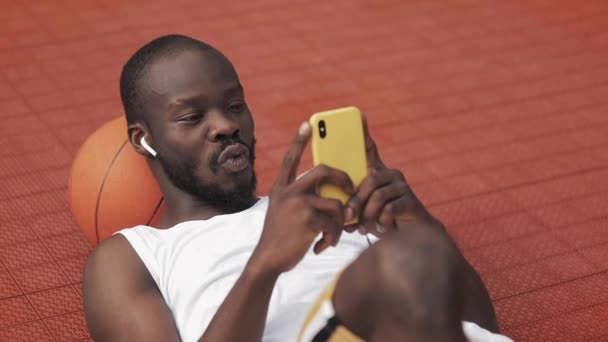 Afro American Guy στα ακουστικά ξαπλωμένος στο έδαφος με μπάλα κάτω από το κεφάλι του, χρησιμοποιώντας Smartphone του, χαμογελώντας, αναζητούν κατάπληκτος και λέγοντας Wow στο γήπεδο μπάσκετ. Υγιής τρόπος ζωής και Αθλητισμού Concept. — Αρχείο Βίντεο