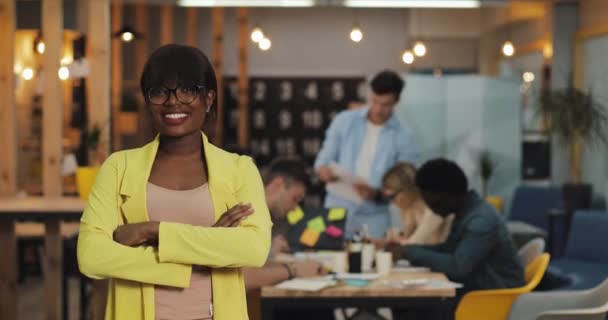 Αφροαμερικανίδα επιχειρηματίας που στέκεται στο σύγχρονο γραφείο, χαμογελώντας και κοιτάζοντας την κάμερα. Οι εργαζόμενοι στο θολό φόντο. — Αρχείο Βίντεο