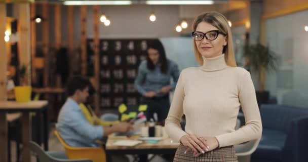 Портрет молодої успішної бізнес-леді зі стильними окулярами, що дивляться на камеру, що стоїть в офісному робочому просторі. Працюючі люди на розмитому тлі . — стокове відео