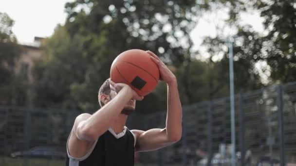 Vista lateral do jovem homem branco musculoso de uniforme preto com cara séria jogando bola em uma cesta, treinando na quadra de basquete de rua Estilo de vida saudável e conceito de esporte . — Vídeo de Stock