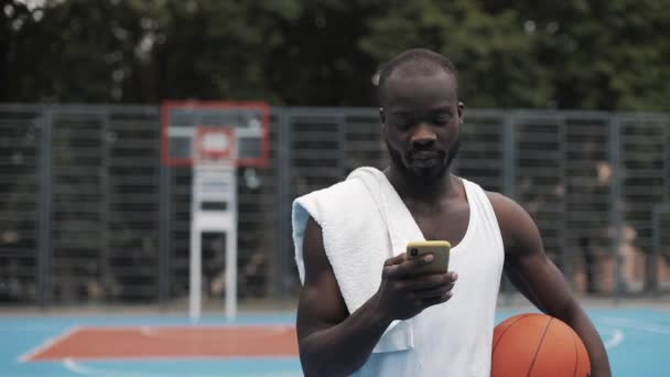 Young Muscly Serious Afro - American Guy in White Singlet Holding Ball em uma mão e usando seu Smartphone com outros Walking at Street Basketbal Court. Estilo de vida saudável e conceito de esporte . — Vídeo de Stock