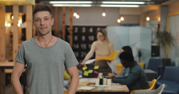 Retrato de un joven hombre de negocios sonriente y exitoso parado en una oficina moderna. Gente trabajadora en el fondo borroso . — Vídeo de stock