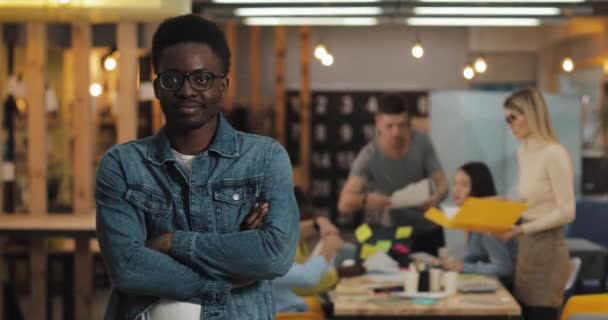 멋진 안경을 쓴 젊은 흑인웃는 사업가가 교차 된 팔로 현대식 사무실에 서 있는 사진. 흐릿 한 배경 위에서 일하는 사람들. — 비디오
