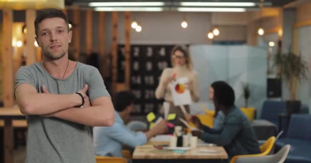 교차 팔을 가진 현대 사무실에 서 있는 젊은 웃는 성공적인 사업가의 초상화. 배경 흐리게 작업 하는 사람들. — 비디오