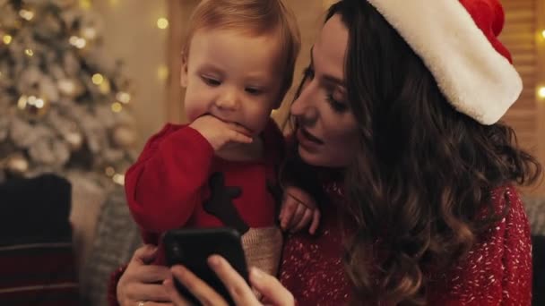 かわいい赤ちゃんと一緒にソファに座っているサンタの帽子をかぶっている若い美しい母の終わり暖かく居心地の良い家の雰囲気でクリスマスツリーの近くで幸せな笑顔を見てビデオ通話をする — ストック動画