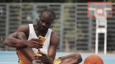 Kulaklıklı Genç Kaslı Afro Amerikalı Adam 'ın Yakın Çekimi, Akıllı Telefonunu kullanarak, Ekranı Scrooling ve Sokak Basketbol Sahasında Gülümseyerek. Sağlıklı Yaşam Tarzı ve Spor Konsepti.