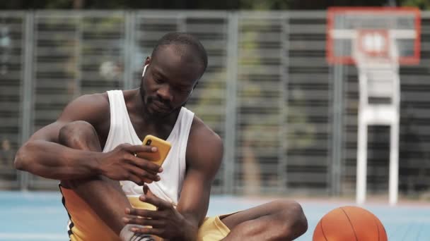 Nahaufnahme eines jungen muskulösen Afroamerikaners mit Kopfhörern, der auf dem Boden sitzt, sein Smartphone benutzt, den Bildschirm scrollt und auf dem Street-Basketballfeld lächelt. gesunder Lebensstil und Sportkonzept. — Stockvideo