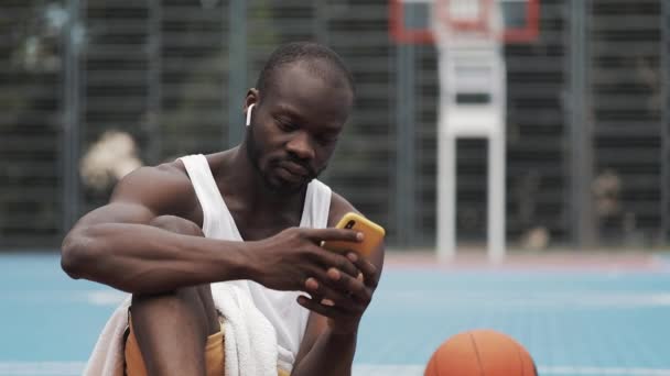 穿着耳机坐在地上的年轻的非洲裔美国人，用他的智能手机，在街头篮球场微笑。 健康的生活方式。 技术与体育概念. — 图库视频影像