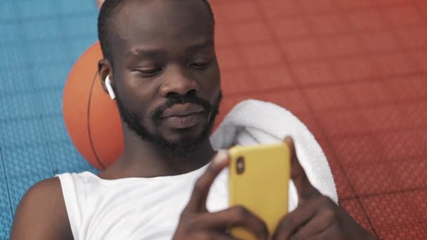 Close Up of Afro American Guy in Earphones Deitado no chão com bola sob sua cabeça, Usando seu Smartphone, Olhando Surpreso e dizendo WoW no Basketball Court. Conceito de desporto . — Vídeo de Stock