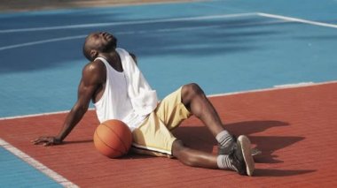 Genç, kaslı, Afro-Amerikan adam yerde oturuyor, yorgun görünüyor, havluyla yüzünü siliyor ve sokak basketbol sahasında oynamaya devam ediyor..