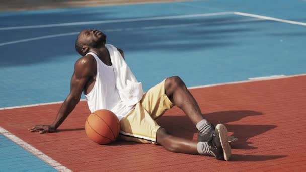 Young Musculy Handsome Afro American Guy Sitting on the Ground, Looking Tired, Wiping Face with a Towel Standing Up and Continue to Play at Street Basketball Court. — Stock Footage Young Musculy Handsome Afro American Guy Sitting on the Ground, Looking Tired, Wiping Face with a Towel Standing Up and Continue to Play at Street Basketball Court. Stock Video