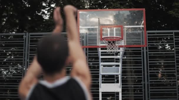 Indietro Veduta del giovane ragazzo caucasico muscoloso in uniforme nera che lancia palla in un cestino e segna, allenandosi al campo da basket di strada. Stile di vita sano e concetto di sport . — Video Stock