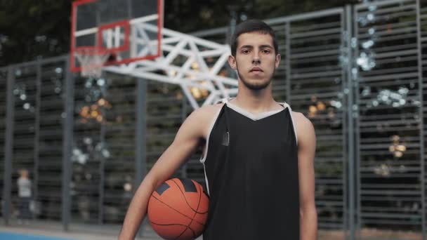 Porträt eines jungen schlanken, ernsthaften kaukasischen Typen in schwarzem Hemd, der Ball am Basketballfeld hält und in die Kamera blickt. gesunder Lebensstil und Sportkonzept. — Stockvideo
