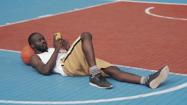 Junger muskulöser, gutaussehender afroamerikanischer Typ, der mit Ball unter dem Kopf auf dem Boden liegt und sein Smartphone auf dem städtischen Basketballplatz benutzt. gesunder Lebensstil und Sportkonzept. — Stockvideo