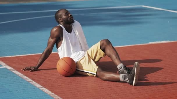 Joven Musculy Guapo Afro-Americano sentado en el suelo, buscando cansado, limpiando la cara con una toalla afuera en la cancha de baloncesto de deportes urbanos. Concepto de estilo de vida saludable y deporte . — Vídeos de Stock