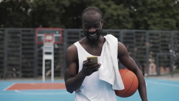 Young Muscly Smiling Afro - American Guy in White Singlet Holding Ball in one hand and Using his Smartphone with other Walking at Street Basketbal Court. Healthy Lifestyle and Sport Concept. — Stock Video