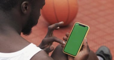 Basketbol sahasında otururken Smartphone, Touching and Scrooling the Screen 'ini kullanan Afro-Amerikan Genç Sakallı Adam' ın Arka Tarafı. Spor ve Teknoloji Konsepti. Yeşil Ekran.