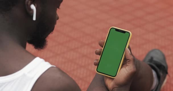 Back Side View of Young Bearded Afro American Guy Wearing earphones Using his Smartphone, Scrooling the Screen while Sitting at Basketball Sport Ground. Концепция спорта и технологий. Зеленый экран . — стоковое видео