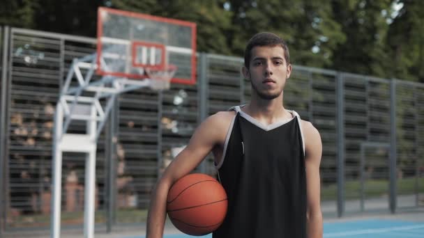 Nahaufnahme Porträt eines jungen schlanken, kaukasischen Basketballspielers in schwarzem Hemd, der Ball am Basketballfeld hält und in die Kamera blickt. gesunder Lebensstil und Sportkonzept. — Stockvideo