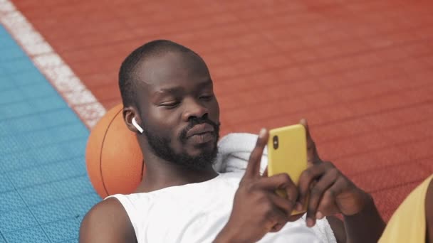 Afro American Guy in Earphones Lying on the Ground with Ball under his Head, Using his Smartphone, Scrooling the Screen at Basketball Court. Mode de vie sain et concept sportif . — Video