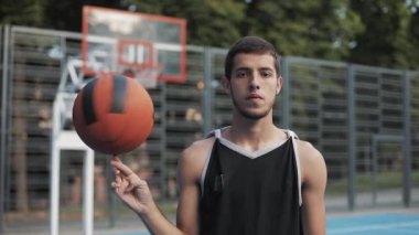 Beyaz tenli genç basketbolcunun kapanışı, Siyah Singlet giyen, topu parmağında döndüren sokak basketbol sahasında duran ve kameraya bakan. Sağlıklı Yaşam Tarzı ve Spor Konsepti.