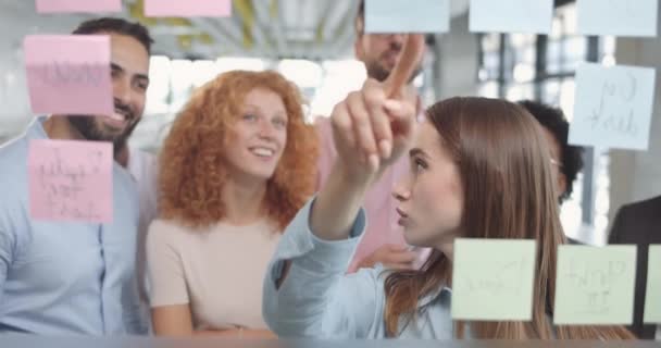 Mujer joven aprendiz hablando con colegas sobre la nueva estrategia del proyecto y la sustitución de notas adhesivas. Trabajadores de oficina compartiendo ideas en reuniones corporativas y utilizando paneles de vidrio . — Vídeos de Stock