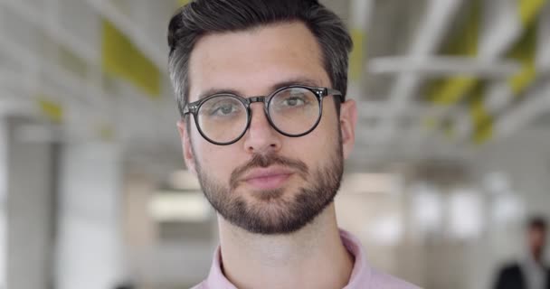 Primer plano del hombre caucásico barbudo en los años 30 mirando a la cámara y parpadeando. Retrato de un tipo guapo con cara seria. Fondo de oficina moderno borroso. Concepto de personas . — Vídeo de stock