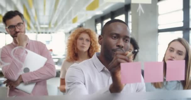 Hombre afroamericano líder del equipo hablando con colegas y escribiendo en tablero de cristal. Jefe de empresa explicando estrategia financiera a sus subordinados en el espacio de trabajo moderno . — Vídeo de stock