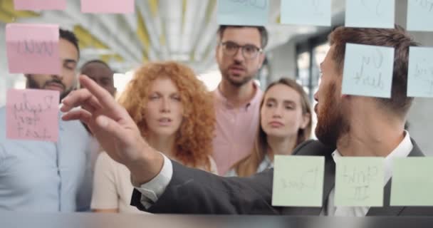 Grupo de trabajadores masculinos y femeninos que tienen animada discusión cerca de tablero de vidrio con notas adhesivas. Coach de negocios masculino en los años 30 de pie y hablando con el equipo multiétnico en la formación corporativa . — Vídeo de stock