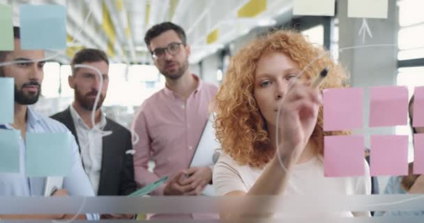 Közelkép egy női irodai munkásról, aki üvegtáblát használ, és javaslatot tesz a problémamegoldásra a munkatársaknak. Különböző munkatársak vállalati találkozóval és üvegfal mellett ragacsos megjegyzésekkel. — Stock videók