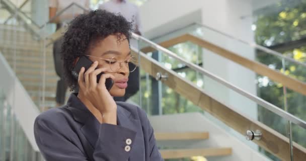 Nahaufnahme einer schönen Büroleiterin, die am Handy spricht und lächelt. junge afrikanische Geschäftsfrau unterhält sich auf ihrem Smartphone und steht im Büro. — Stockvideo