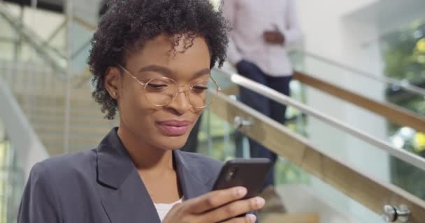 Primer plano de la mujer de negocios africana en gafas usando el teléfono y sonriendo en el moderno edificio de oficinas. Trabajadora de oficina navegando por la red, deslizando, mensajes de texto en el teléfono inteligente. Concepto de personas, tecnología . — Vídeos de Stock