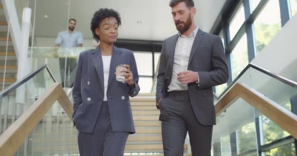 Handsome businessman and his attractive african coworker walking downstairs and chatting. Young woman manager with coffee cup and her male colleague descending office stairs and talking. — Stock Video