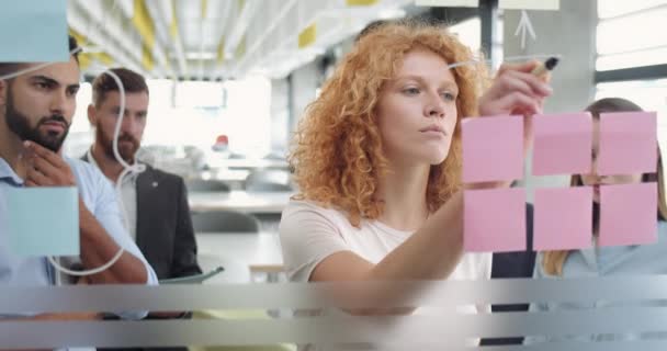 Mulher ruiva jovem sugerindo colegas de escritório sua visão conceitual ao desenhar na parede de vidro. Equipe jovem corporativa que está perto de placa de vidro e fazendo estratégia, planejamento, brainstorming . — Vídeo de Stock