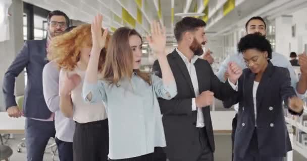 Lucrătorii de birou entuziasmați dansează, zâmbesc, se simt bine în spațiul de lucru modern. Vedere din față a echipei de afaceri fericite care se bucură de succesul startup-ului. Conceptul de victorie în echipă, internaționalitate . — Videoclip de stoc