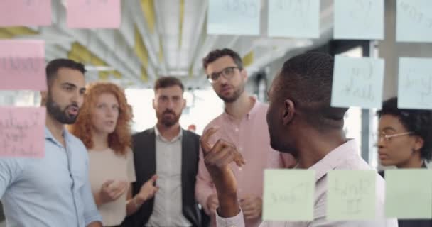 Aconselhamento de membros da equipe affrican masculino com colegas e substituindo notas pegajosas. Grupo de trabalhadores de escritório multi étnicos de pé e brainstorming em reunião de escritório usando placa de vidro . — Vídeo de Stock