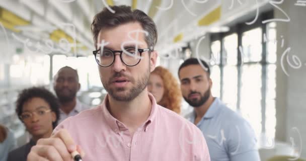 Trabajador de oficina masculino en gafas escribiendo en tablero de cristal y explicando el concepto a los miembros del equipo. Primer plano del hombre barbudo guapo de pie cerca de la pared de vidrio en la conferencia corporativa . — Vídeo de stock