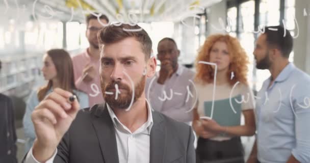 Biurowiec z brodą, pisze formułę na szklanej tablicy i myśli. Grupa mężczyzn i kobiet stojących i patrzących na szefa w pobliżu szklanej ściany na spotkaniu korporacyjnym. — Wideo stockowe