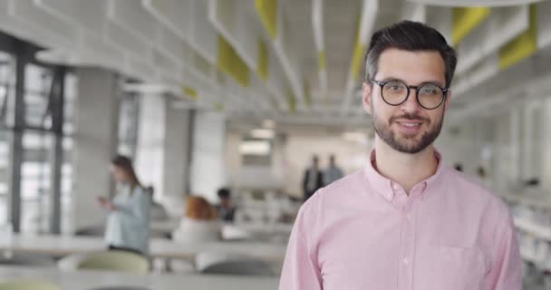 カメラと笑顔に頭を回すメガネで陽気な男性オフィスの労働者。現代のコワーキング・ゾーンに立つハッピー・ベアード・マンのスタートアップ・ビジネスオーナー。仕事、キャリア、人々の概念. — ストック動画