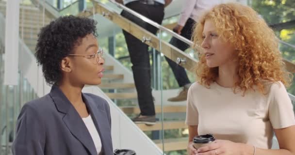 Duas mulheres de raça diversificada trabalhadores de escritório em pé no corredor do escritório e conversando. Close up de jovens colegas de trabalho bem sucedidos bebendo cofee, tendo conversa agradável e rindo . — Vídeo de Stock